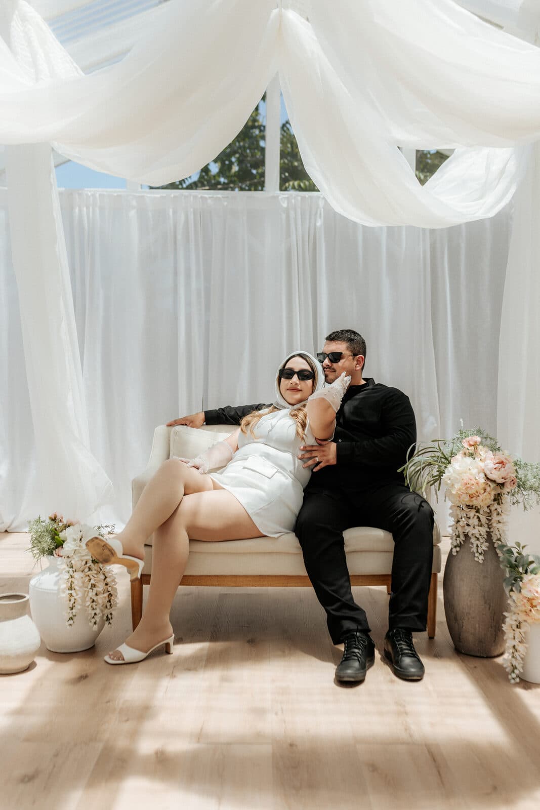 Stylish couple seated in a white draped cabana with florals