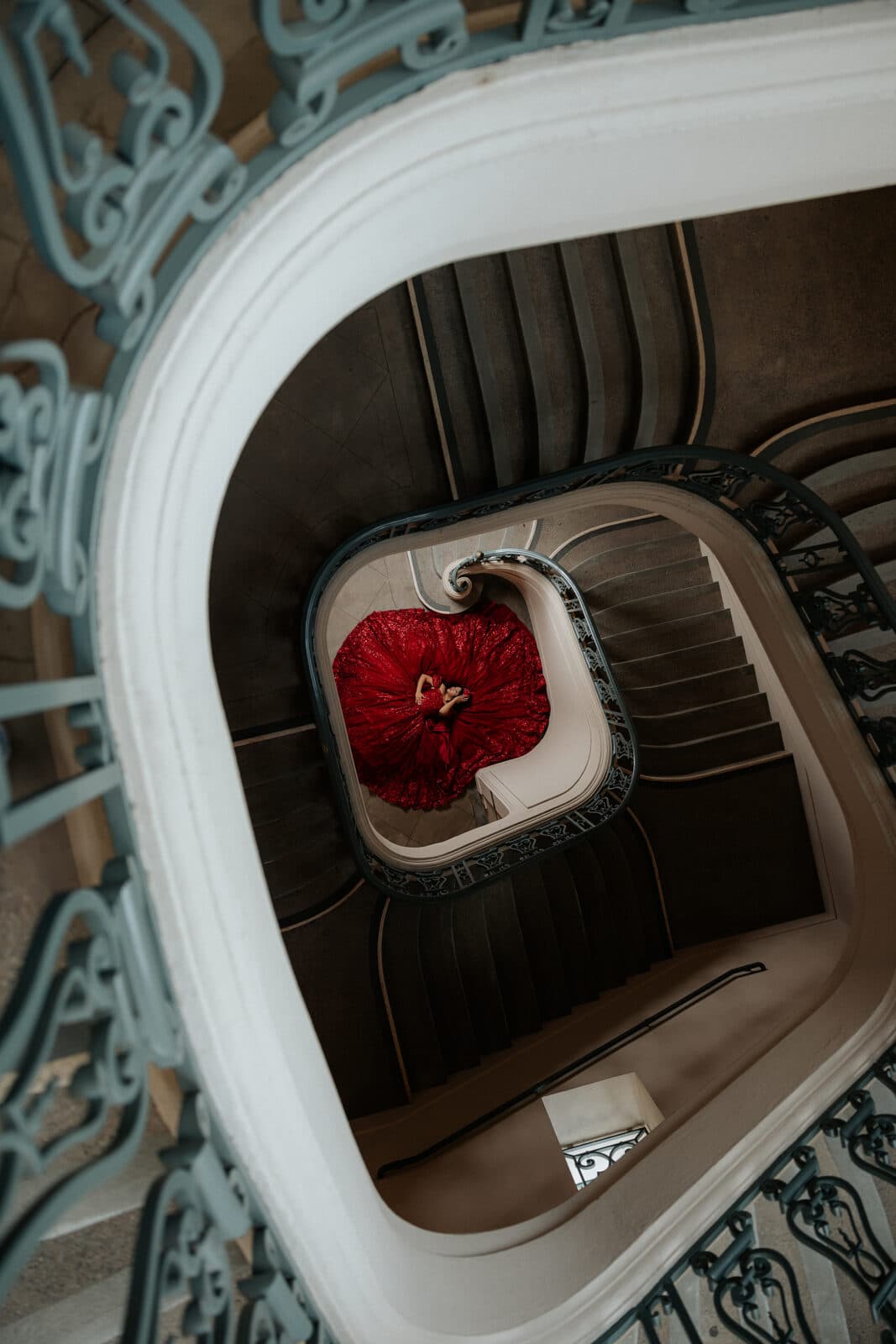 Quinceañera in a red gown photographed from above through an ornate spiral staircase